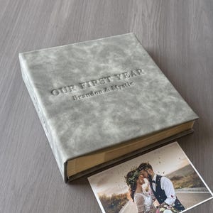 May include: A grey, textured photo album with the embossed text "OUR FIRST YEAR" and "Brandon & Mystic." A photograph of a couple kissing is next to the album. The album is a square shape.