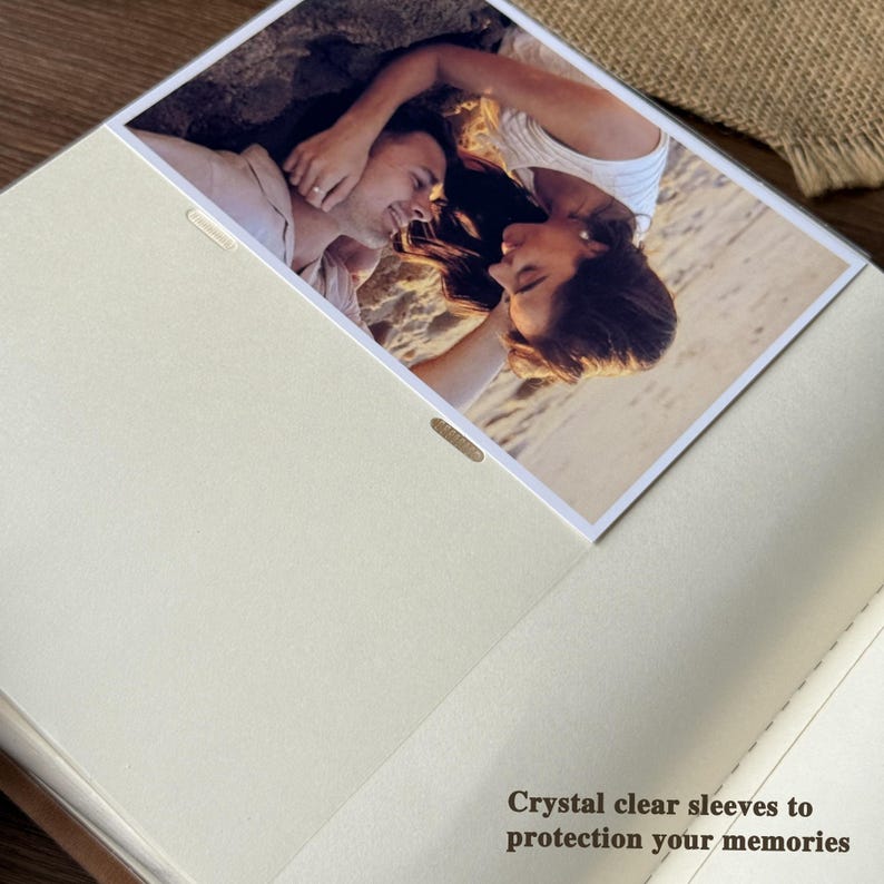 Peut inclure: Un album photo beige ouvert avec une photo d'un couple. L'album a des pochettes transparentes et le texte "Crystal clear sleeves to protection your memories". La photo montre un couple sur une plage.