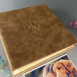 Puede incluir: Un álbum de fotos marrón de cuero sintético con una huella de pata en relieve y el nombre "Don". El álbum está abierto, revelando páginas color crema. Se ve una fotografía de un golden retriever.
