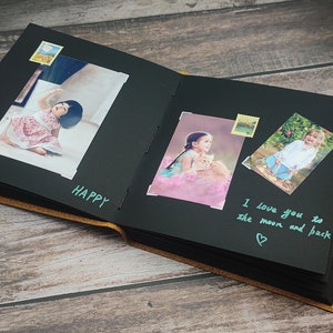 Puede incluir: &Aacute;lbum de fotos negro con una cubierta de cuero marr&oacute;n. El &aacute;lbum est&aacute; abierto para revelar p&aacute;ginas negras con fotos de ni&ntilde;os y mensajes escritos a mano. Los mensajes incluyen "Happy" y "Te amo hasta la luna y de regreso".