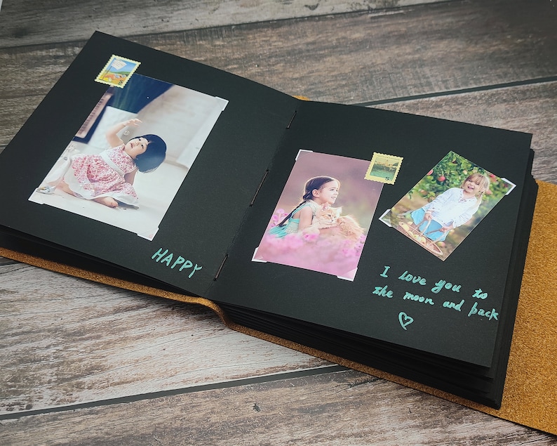 Puede incluir: &Aacute;lbum de fotos negro con una cubierta de cuero marr&oacute;n. El &aacute;lbum est&aacute; abierto para revelar p&aacute;ginas negras con fotos de ni&ntilde;os y mensajes escritos a mano. Los mensajes incluyen "Happy" y "Te amo hasta la luna y de regreso".