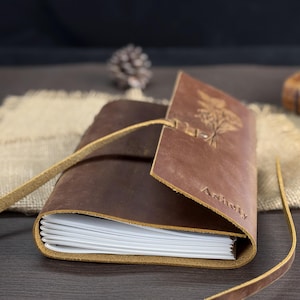 May include: A brown leather journal with a strap closure. The journal has a debossed botanical design and the name "Ashely" on the cover. The pages are white and the journal is open, revealing the interior pages.