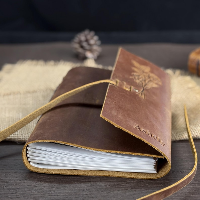 May include: A brown leather journal with a strap closure. The journal has a debossed botanical design and the name "Ashely" on the cover. The pages are white and the journal is open, revealing the interior pages.