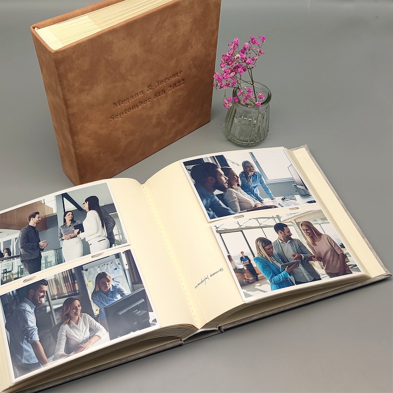 Peut inclure: Un album photo en daim marron avec les noms "Megann & Jeremy" et la date "September 5th 2022" estampill&eacute;s sur la couverture. L'album est ouvert, affichant plusieurs photographies. Un petit vase avec des fleurs roses est &eacute;galement pr&eacute;sent.