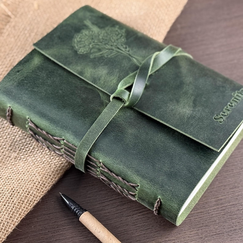 May include: Green leather journal with a floral design embossed on the cover. The journal has a wraparound strap closure and stitched binding. The word "Serenity" is embossed on the cover. A pen rests beside the journal.