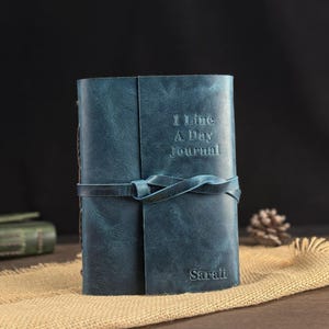 May include: A blue leather journal with the embossed text "1 Line A Day Journal" and the name "Sarah". The journal is secured with a matching leather tie. The journal is sitting on a burlap surface.