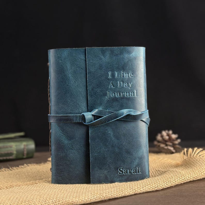 May include: A blue leather journal with the embossed text "1 Line A Day Journal" and the name "Sarah". The journal is secured with a matching leather tie. The journal is sitting on a burlap surface.