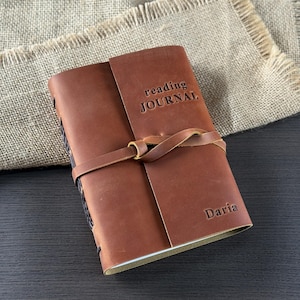 Peut inclure: Un journal de lecture en cuir marron avec les mots "reading JOURNAL" en relief sur la couverture. Le journal a une fermeture à lanière en cuir et le nom "Daria" est estampé en bas à droite.