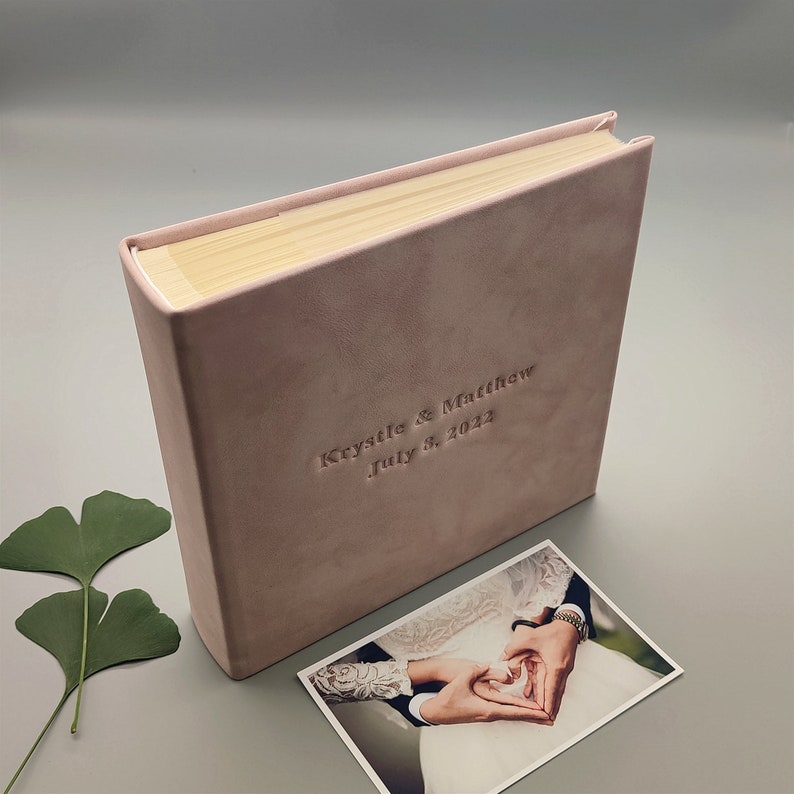 Peut inclure: Un album de mariage rose p&acirc;le avec l'inscription en relief "Krystle & Matthew July 3, 2022". Une photo d'un couple se tenant la main est &agrave; c&ocirc;t&eacute; de l'album. Les pages de l'album sont visibles en haut.