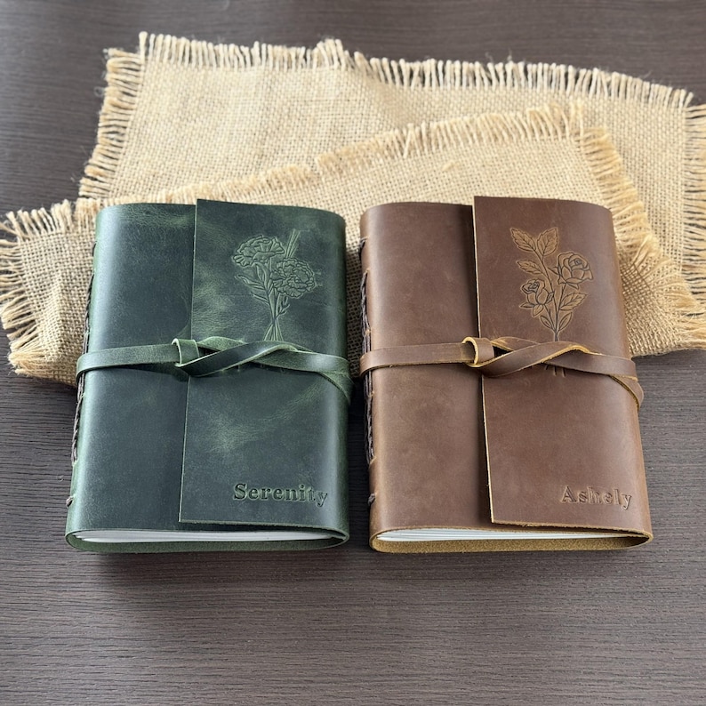 May include: Two leather journals, one dark green and one brown, with embossed floral designs. The green journal has the word "Serenity" embossed on the cover, while the brown journal has "Ashley". Both have a leather strap closure.