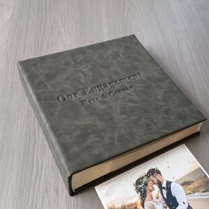May include: A grey leather photo album with the embossed text "Our Engagement Kate & Connor." A photograph of a kissing couple is partially visible. The album has a textured surface and a classic design.