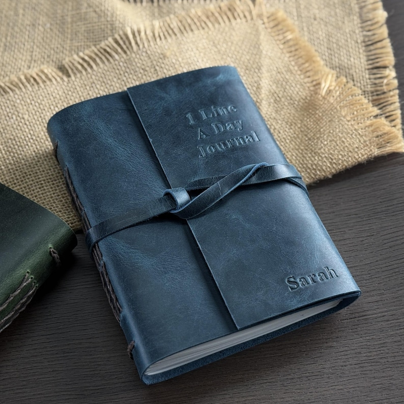 May include: A blue leather journal with the text "1 Line A Day Journal" embossed on the cover. The journal is secured with a leather strap and features the name "Sarah" embossed on the bottom right corner.