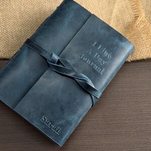 May include: A blue leather journal with the text "1 Line A Day Journal" embossed on the cover. The journal is closed with a leather strap tied in a knot. The name "Sarah" is embossed on the bottom corner.