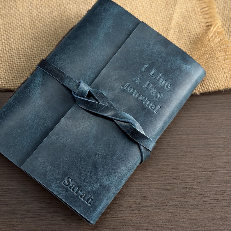 May include: A blue leather journal with the text "1 Line A Day Journal" embossed on the cover. The journal is closed with a leather strap tied in a knot. The name "Sarah" is embossed on the bottom corner.
