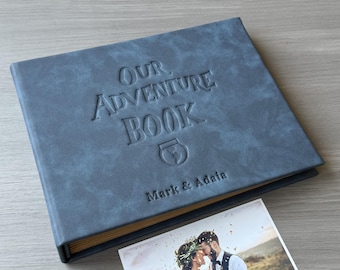 May include: A blue photo album with the embossed text "OUR ADVENTURE BOOK" and the names "Mark & Adaia". A photograph of a couple is next to the album. The album has a textured cover.