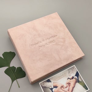 Peut inclure: Un album photo de mariage rose p&acirc;le avec le texte en relief "Krystle & Matthew July 8, 2022." Une photo des mains d'un couple formant un c&oelig;ur est visible. L'album est sur une surface grise avec une feuille verte.