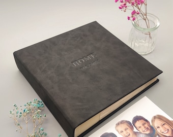 May include: A dark gray photo album with the word "HOME" and "VOLUME 1" embossed on the cover. The album is rectangular and appears to be made of textured material. A small glass vase with pink flowers and a photo of a family are also in the image.