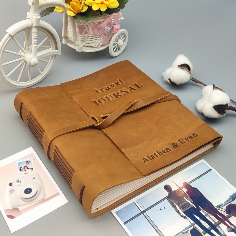 Puede incluir: Un diario de viaje de cuero marr&oacute;n con una correa y las palabras "travel journal" grabadas en la parte delantera. El diario est&aacute; personalizado con los nombres "Alathea & Evan".