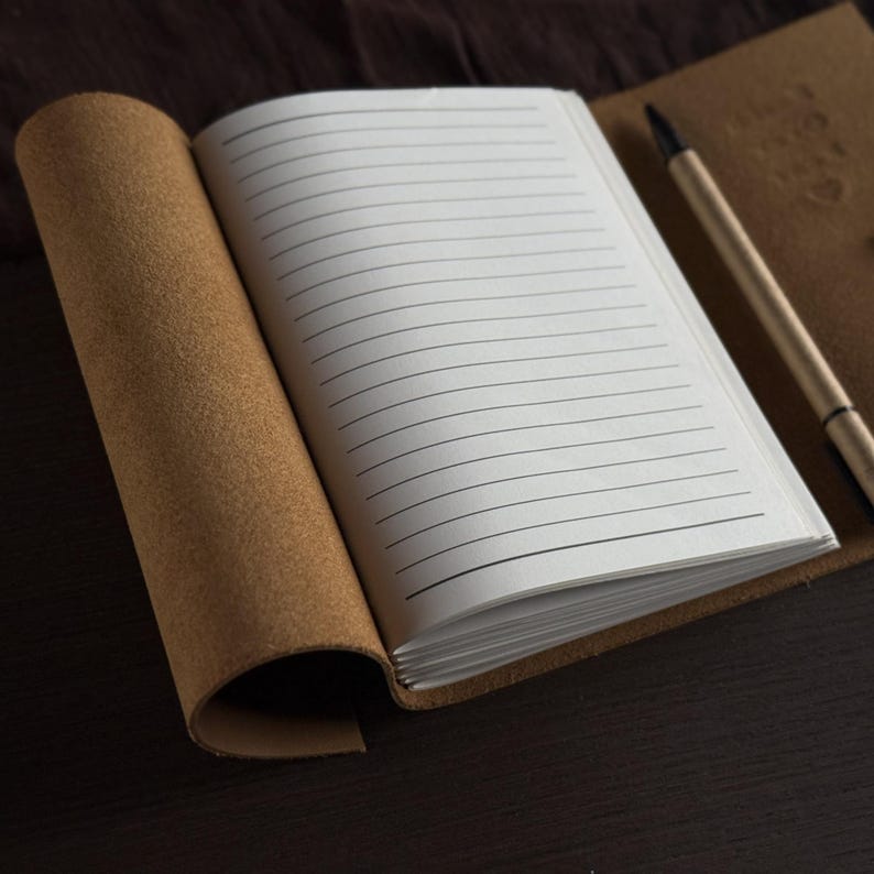 May include: Open, lined notebook with a brown leather cover and a matching pen. The pages are white with black lines, and the notebook is open to a blank page. The pen is silver and brown.