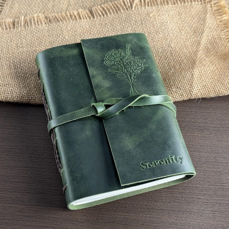 May include: Green leather journal with a floral design embossed on the cover. The journal has a wraparound tie closure and the word "Serenity" embossed on the front. The journal is closed and resting on a wooden surface.