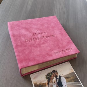 May include: A pink photo album with the embossed text "Abby's Bridal Shower" and "July 22 2025". A photograph of a couple is next to the album. The album has a soft, textured cover.