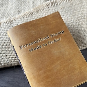 May include: A tan leather journal with the embossed text "Personalized Stamp Made to Order." The journal has a stitched spine and is set against a neutral fabric background.