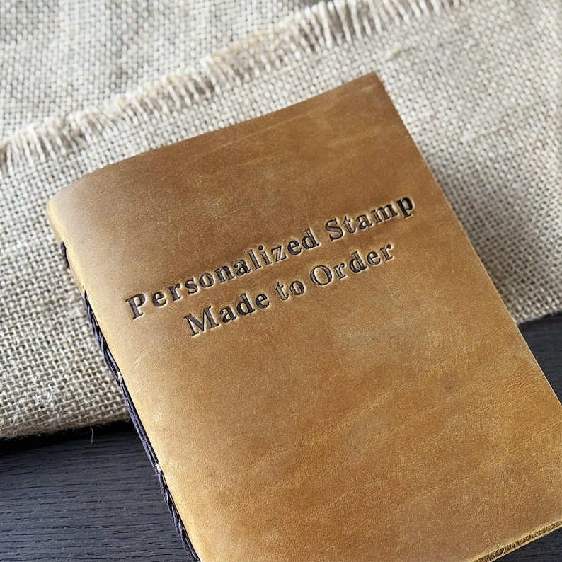 May include: A tan leather journal with the embossed text "Personalized Stamp Made to Order." The journal has a stitched spine and is set against a neutral fabric background.