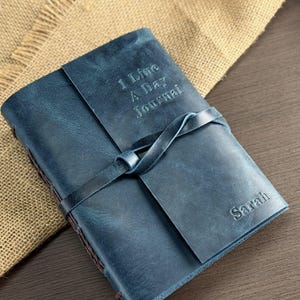 May include: A blue leather journal with the text "1 Line A Day Journal" embossed on the cover. The journal is closed with a leather strap and the name "Sarah" is embossed on the bottom right corner.
