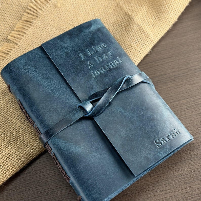 May include: A blue leather journal with the text "1 Line A Day Journal" embossed on the cover. The journal is closed with a leather strap and the name "Sarah" is embossed on the bottom right corner.