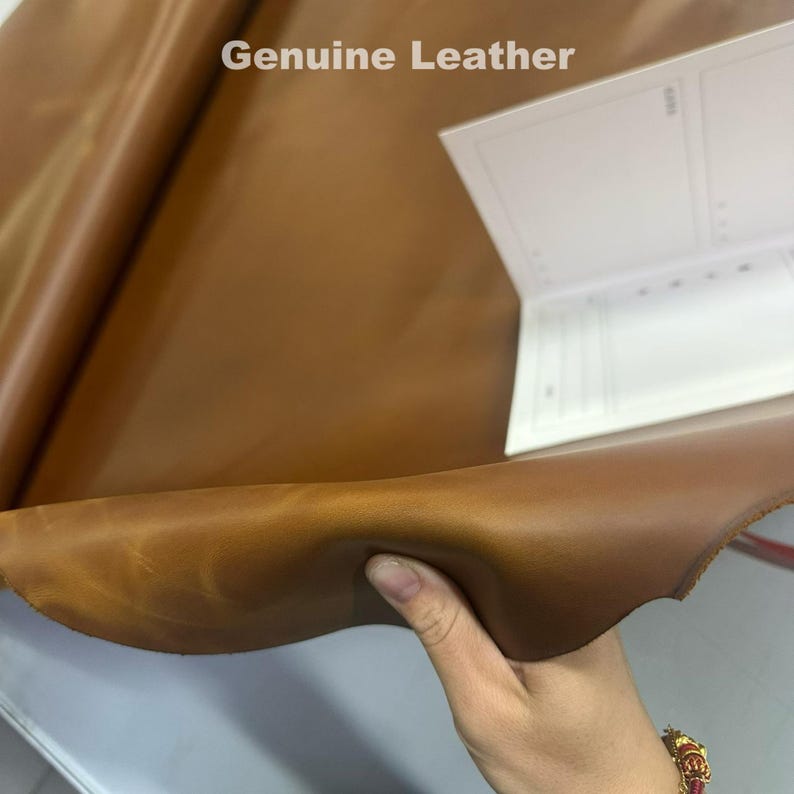 May include: A close-up of a sheet of brown genuine leather. The leather is folded and held by a hand. The text "Genuine Leather" is visible in the upper left corner. A white notepad is in the background.