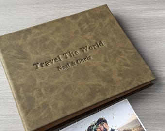 Könnte beinhalten: Ein strukturiertes, olivgrünes Fotoalbum mit dem geprägten Text "Travel The World" und "Neal & Chris". Ein gedrucktes Foto eines sich küssenden Paares ist neben dem Album zu sehen. Das Album ist rechteckig und scheint ledergebunden zu sein. Etwa 30 cm lang.
