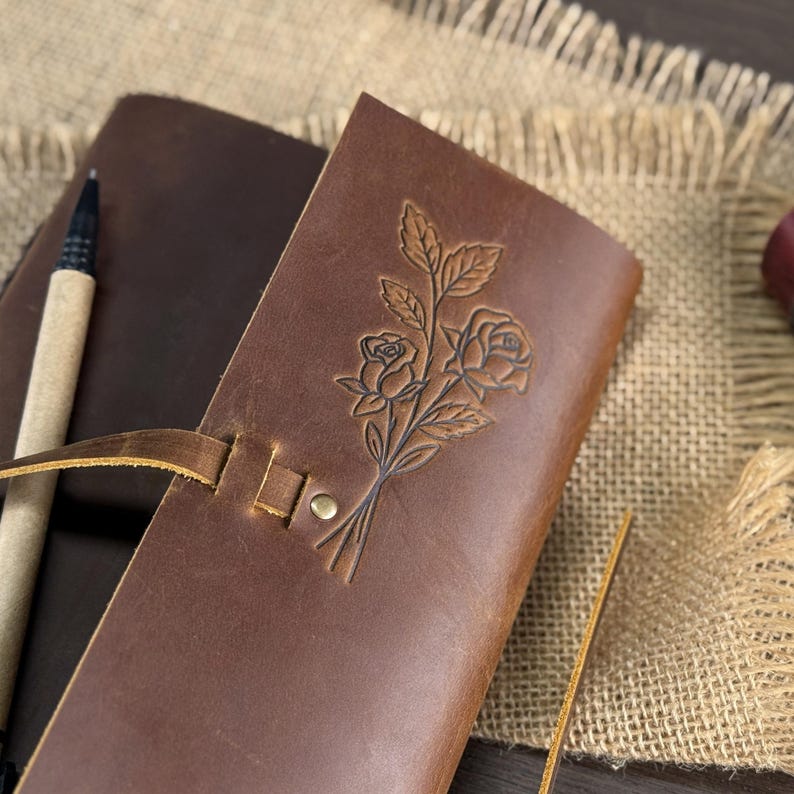 May include: A brown leather journal with a floral rose design embossed on the cover. The journal is closed with a leather strap and a brass button. A pen rests beside the journal on a burlap surface.