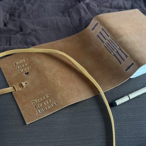 May include: A tan leather journal with the embossed text "Faith Over Fear" and "Steve's Prayer Journal." The journal is open, revealing white pages, and has a leather strap closure. A pen rests beside the journal.