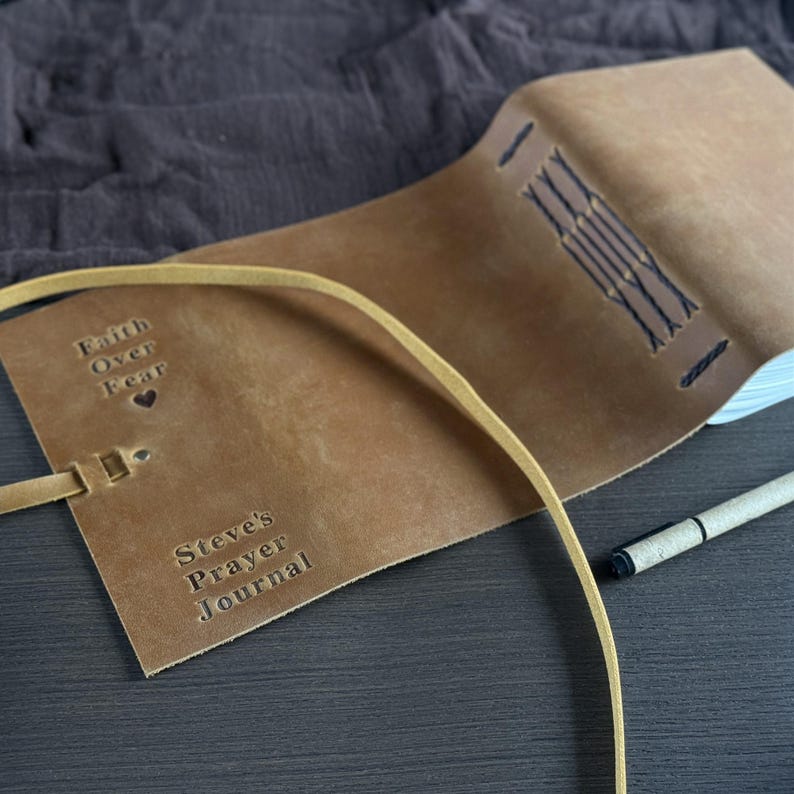 May include: A tan leather journal with the embossed text "Faith Over Fear" and "Steve's Prayer Journal." The journal is open, revealing white pages, and has a leather strap closure. A pen rests beside the journal.