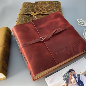 Puede incluir: Un &aacute;lbum de fotos de cuero burdeos con una correa de cuero marr&oacute;n y el texto "Emma & Matthew 2024" grabado en la parte delantera. El &aacute;lbum est&aacute; cerrado y se encuentra sobre una mesa con una foto de boda en el fondo.