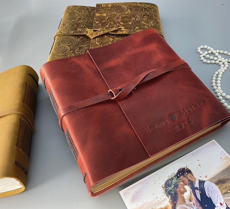 Puede incluir: Un &aacute;lbum de fotos de cuero burdeos con una correa de cuero marr&oacute;n y el texto "Emma & Matthew 2024" grabado en la parte delantera. El &aacute;lbum est&aacute; cerrado y se encuentra sobre una mesa con una foto de boda en el fondo.