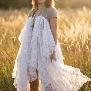 Puede incluir: Una persona en un campo sostiene un vestido de encaje blanco sobre una parte superior gris. El vestido presenta intrincados patrones florales y un dobladillo asimétrico. También se ve un collar de perlas y hierba alta.
