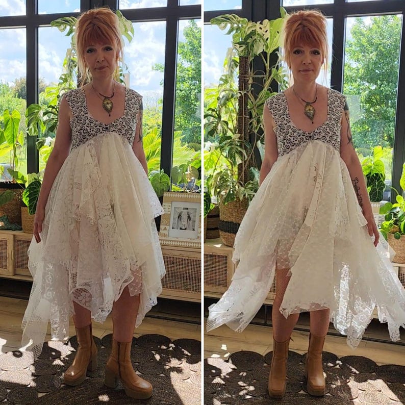 May include: A white lace dress with a unique asymmetrical hemline. The dress features a detailed bodice with a black and white pattern and a flowing skirt. The model is wearing brown platform boots. The background includes lush greenery and natural light.