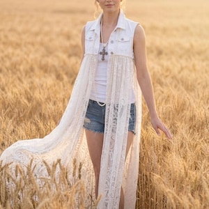 Puede incluir: Un chaleco vaquero blanco sin mangas con una larga superposición de encaje blanco. El chaleco se combina con pantalones cortos de mezclilla y una camiseta blanca. Un collar de perlas con un colgante de cruz completa el look. El fondo es un campo de trigo dorado.