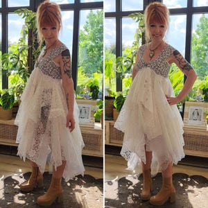 May include: A white lace dress with a high-low hemline and a patterned bodice. The dress features a layered skirt and is paired with tan platform boots. The background includes a window with plants and a wooden cabinet.