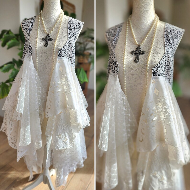 May include: A cream-colored lace dress with a black and white patterned vest. A pearl necklace with a cross pendant hangs over the dress. The dress is displayed on a mannequin.