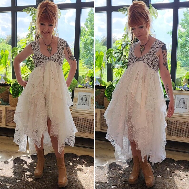 May include: A white lace dress with a high-low hemline. The dress features a detailed bodice with a patterned design and a flowing skirt. The model is wearing brown platform boots. The background includes a large window and greenery.