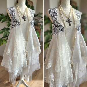 May include: A white lace dress with a layered design and a black and white patterned vest. The dress is adorned with a pearl necklace and a cross pendant. The dress is displayed on a mannequin.