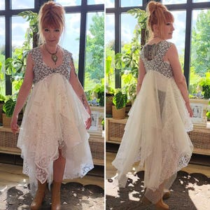 May include: A sleeveless, cream-colored dress with a high-low hemline. The bodice is patterned with a black and white design, and the skirt features layers of lace. The dress is worn with brown boots.