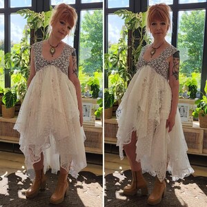 May include: A white lace dress with a high-low hemline. The dress features a detailed bodice with a black and white pattern and a flowing skirt. The model is wearing brown platform boots. The background includes lush green plants and natural light.