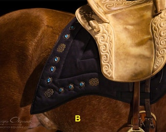 May include: A close-up of a brown horse with a black saddle pad decorated with blue gemstones and gold accents. The saddle pad is under a tan leather saddle with intricate stitching.