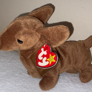 Op de afbeelding: Bruin pluche speelgoedhondje met een rood Beanie Baby-label waarop "Beanie Babies" staat en een ster.