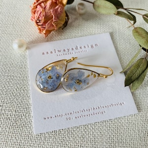 May include: Gold teardrop earrings with blue pressed flowers and gold leaf accents. The earrings are displayed on a white card with the text "asalwaysdesign handmade jewelry" and website information.