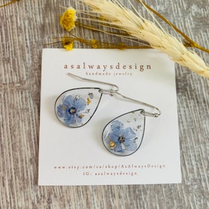 May include: A pair of silver teardrop earrings with a blue flower pressed inside each teardrop. The earrings are handmade and feature gold leaf accents. The earrings are displayed on a white card with the text "asalwaysdesign handmade jewelry" and the website address "www.etsy.com/ca/shop/AsAlwaysDesign" and Instagram handle "IG: asalwaysdesign".