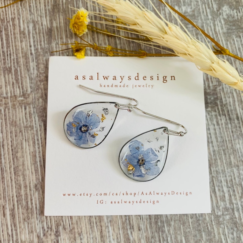 May include: A pair of silver teardrop earrings with a blue flower pressed inside each teardrop. The earrings are handmade and feature gold leaf accents. The earrings are displayed on a white card with the text "asalwaysdesign handmade jewelry" and the website address "www.etsy.com/ca/shop/AsAlwaysDesign" and Instagram handle "IG: asalwaysdesign".